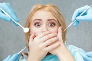 A woman covering her mouth as gloved hands that are holding dentist tools reach out to her