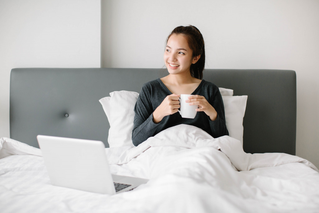 a woman in bed sitting beside a laptop and holding a mug